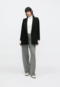 Zwarte oversized blazer met revers, gecombineerd met een witte coltrui en grijze krijtstreepbroek. Schoeisel is wit met zwarte zolen.