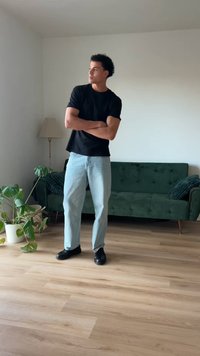 Μαύρο κοντομάνικο μπλουζάκι, ανοιχτό μπλε παντελόνι με ίσια γραμμή και μαύρα παπούτσια. Πράσινος βελούδινος καναπές και φυλλώδες φυτό σε φωτεινό δωμάτιο.