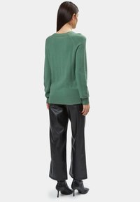 Maglione verde lavorato a maglia con scollo ampio, polsini e orlo a coste, abbinato a pantaloni neri in pelle a gamba larga e stivali con tacco alla caviglia.