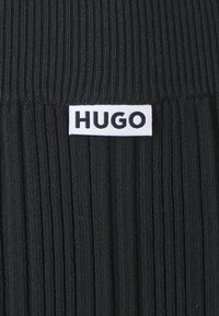 Crna rebrasta tkanina s vertikalnim linijama, s bijelom etiketom koja prikazuje ime brenda "HUGO" u podebljanim crnim slovima.