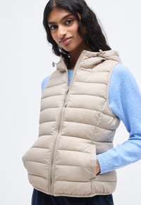 Μπεζ καπιτονέ γιλέκο με κουκούλα, με κάθετα καπιτονέ πάνελ και εμπρός φερμουάρ. Ορατή καπιτονέ υφή και δύο πλαϊνές τσέπες.