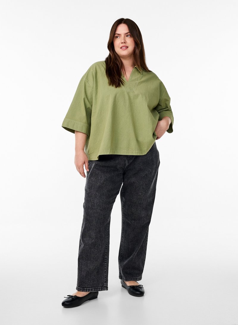 Kvinde med langt brunt hår iført en oversize lysegrøn skjorte, sorte vidde jeans og sorte flade sko, står med den ene hånd i lommen.