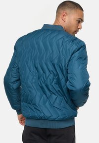 Tirkizna prešana bomber jakna s chevron uzorkom, rebrastim ovratnikom i manžetama. Glatka tekstura i elastični pojas za prilagođeni izgled.