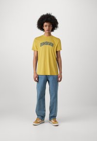 Yellow T-shirt with "QUIKSILVER" graphic in dark green, paired with light blue jeans and beige sneakers featuring brown accents.