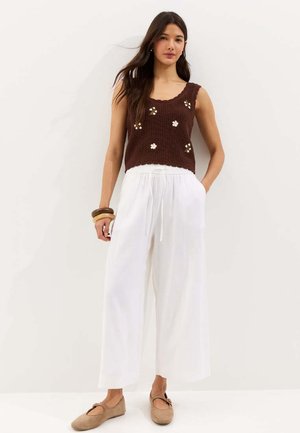 Joven mujer con un top marrón sin mangas de punto con bordado floral, pantalones blancos de pierna ancha, zapatos planos beige y brazaletes de madera, de pie contra una pared blanca.