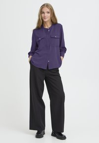 Lillakast nööbivaba särk koos kahe rinna taskuga, pikad varrukad ja ümara kaelusega, kombineeritud mustade laialdaste pükste ja põlvekatega saapatega.