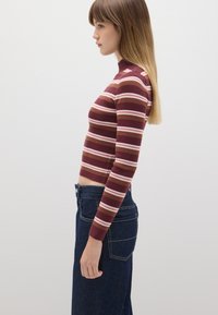 Suéter de cuello alto de color marrón con trenzado y rayas horizontales en rosa y marrón, combinado con jeans de mezclilla oscuros de cintura alta.