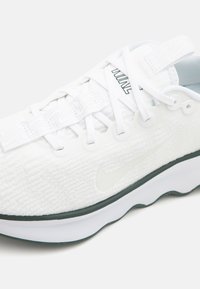 Hvit treningssko med teksturert stoffoberdel, snøring og plastisk hælflik. Har et grønt accent på sålen og Nike-logo.