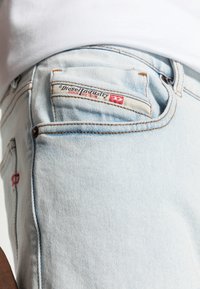 Jean en denim bleu clair avec un patch "Diesel Industry Denim Division" en rouge. Les coutures sont de couleur camel contrastante.