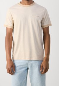 Beige T-shirt med lys tan-ribkant ved krave og manchetter. Blødt bomuldsstof; har et diskret logo på venstre bryst. Pasform: afslappet.