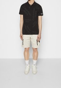 Černé polo tričko s opakujícím se textem "Calvin Klein Jeans", v kombinaci s béžovými cargo šortkami se zapínacími kapsami a bílými teniskami.