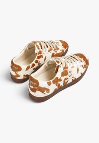 Zapatillas con patrón de piel de vaca en crema y marrón, con un diseño de corte bajo, cierre con cordones y suela de goma marrón.