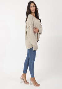 Jersey de punto beige de gran tamaño con puños y dobladillo acanalados, combinado con jeans ajustados azules y sandalias de tacón cuadrado transparente.