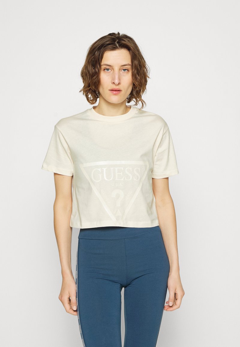 Supjaustyta kreminė medvilninė marškinėliai, su dideliu trikampiu grafiku, kuriame užrašas "GUESS" sidabro spalva ir klausimo ženklas. Suderinti su mėlynomis leggingomis.