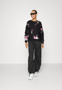 Black sweater with floral embroidery, paired with black cargo pants. Model wears black sunglasses and white sneakers with gray accents.