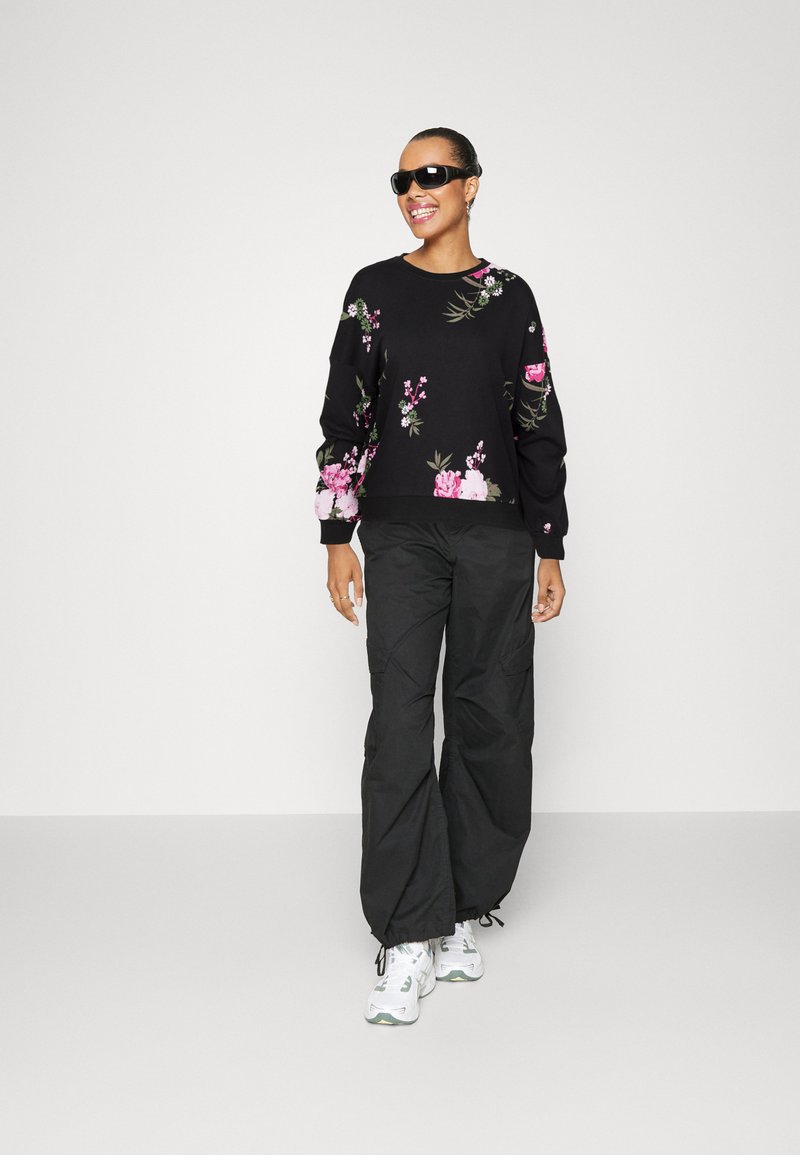 Pull noir avec broderies florales, associé à un pantalon cargo noir. Le mannequin porte des lunettes de soleil noires et des baskets blanches avec des accents gris.