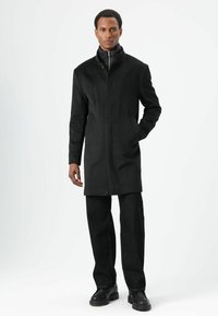 Zwarte lange jas met een hoge kraag en een rits aan de voorkant, gecombineerd met donkere jeans en zwarte schoenen; gladde stof, een getailleerde pasvorm, minimale accenten.