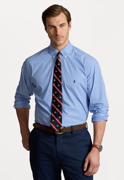Chemise bleu vichy avec col boutonné, associée à une cravate rayée rouge et marine avec un motif d'abeilles. Pantalon marine et ceinture marron.
