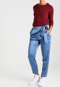 Pull moulant couleur bordeaux associé à un pantalon taille haute en satin bleu clair avec un nœud sur le côté, complété par des baskets blanches.