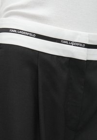 Biely pás so čiernym pruhom a textom "KARL LAGERFELD" nad čiernymi nohavicami a bielym rebrovaným topom.