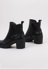 Bottes noires en cuir à la cheville avec un talon épais, fermeture éclair latérale, boucle décorative sur une sangle et semelle texturée.