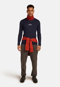 Camicia a maniche lunghe blu navy con collo alto e logo, abbinata a pantaloni grigio scuro e una felpa arancione legata in vita. Scarpe nere.