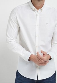 Chemise blanche à boutons avec manches longues, confectionnée dans un tissu léger. Présente un petit logo brodé sur la poitrine et des poignets boutonnés.