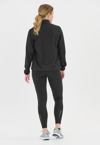 Svart lättviktsjacka med ståkrage, kombinerad med svarta leggings med texturerad detalj och reflexaccent. Silverfärgade löparskor.
