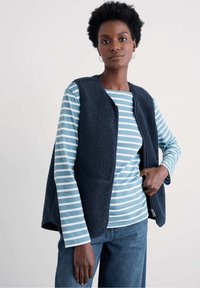 Tumesinine fliisvest tekstuurse viimistlusega, ees tõmblukk ja küljetaskud, kantud hele sinise ja valge triibulise pika varrukaga särgi peal.