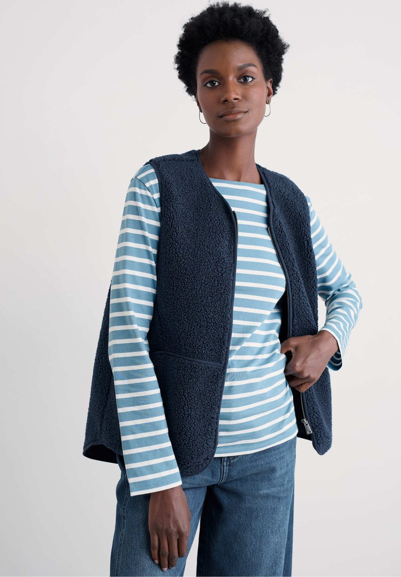 Tumesinine fliisvest tekstuurse viimistlusega, ees tõmblukk ja küljetaskud, kantud hele sinise ja valge triibulise pika varrukaga särgi peal.