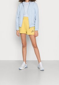 Veste bomber bleu clair, shorts jaunes avec logo, chaussures de sport blanches avec accents bleu clair, t-shirt uni. Textures lisses et coupe décontractée.