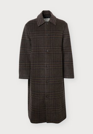 Un mannequin masculin se tient devant un fond uni, vêtu d’un long manteau marron à carreaux, d’un pantalon noir à revers et de chaussures noires.