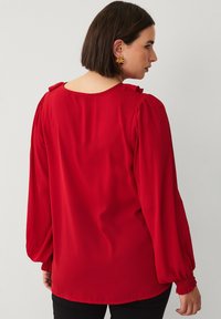 Rode blouse met opgeblazen lange mouwen, voorzien van textuuraccenten op de schouders en elastische manchetten, gemaakt van een gladde stof.