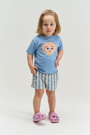 Mladé dievča stojí, má na sebe modré tričko s grafikou škľábajúcej sa mušle, pruhované šortky, ružové topánky a ružovú sponku do vlasov.