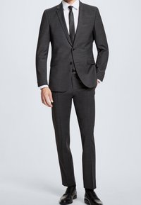 Mörkgrå, skräddarsydd kostym med en strömlinjeformad design, skarpa slag och ett texturerat tyg. Kombinerad med en svart slips och polerade skor.