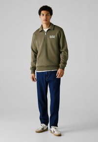 Sudadera con cremallera en verde oliva y puños acanalados, que presenta el logo de "Red Bull Racing". Combinada con jeans azules y zapatillas blancas.