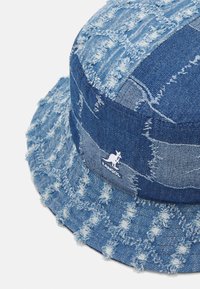 Cappello bucket in denim di diverse tonalità di blu, con orli sfrangiati e design in patchwork, caratterizzato da un logo bianco del canguro di lato.