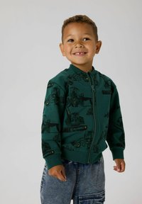 Zelená bundu na zip s vzorem stavebních vozidel, s žebrovaným límcem a manžetami, kombinováno s džínovými šortkami.