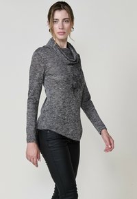Langärmliges Oberteil mit Kapuzenkragen aus strukturiertem grauem Stoff, mit taillierter Passform und asymmetrischem Saum, kombiniert mit schwarzen Hose.