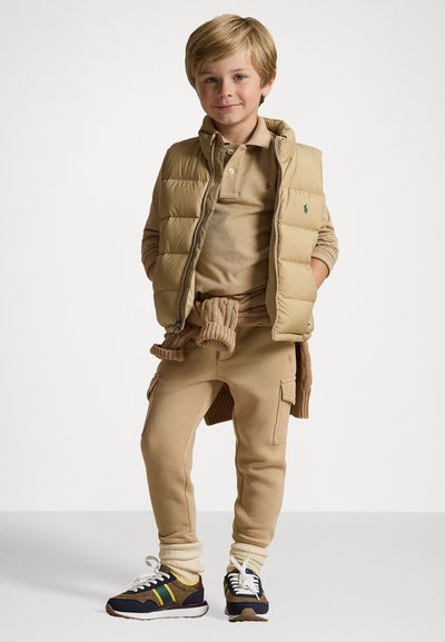 Colete de puffer bege sobre uma polo bege, combinado com calças de fato da carga em tons de bege e sapatilhas com detalhes multicoloridos e atacadores brancos.