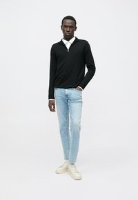 Schwarzer Pullover mit Reißverschluss am Kragen, hellblaue Jeans und cremefarbene Sneakers. Der Pullover hat lange Ärmel und eine taillierte Passform.