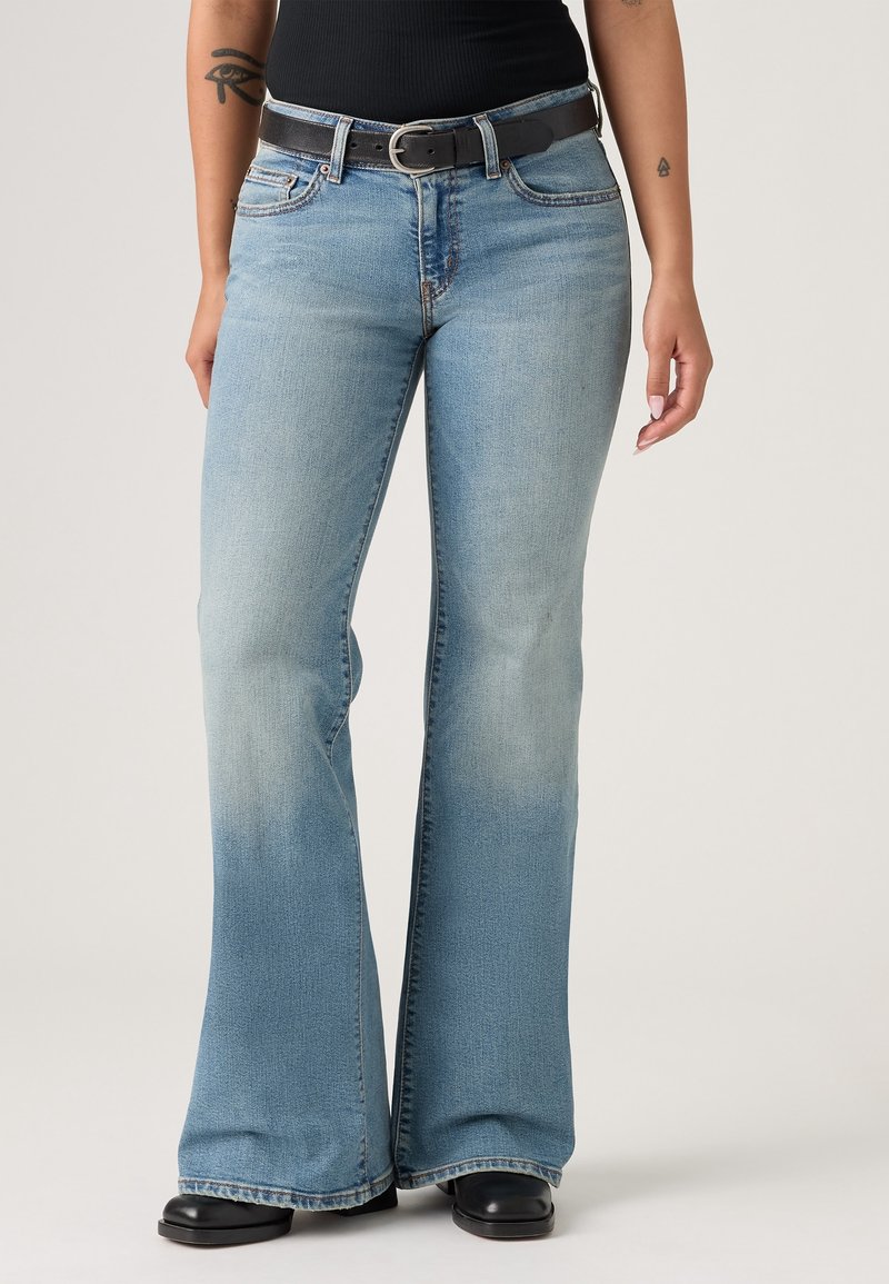 Personne portant un jean évasé bleu clair avec une ceinture noire, un haut noir, des chaussures noires, et des tatouages visibles sur les bras.