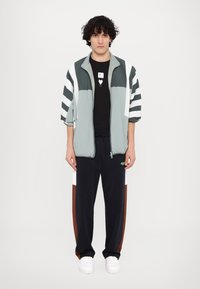 Sporty outfit med en lys grøn lynlåsjakke med sort og hvid stribede ærmer, en sort grafik t-shirt og marineblå sweatsbukser med brune detaljer.