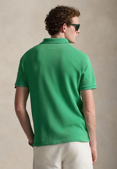 Άνδρας με σγουρά μαλλιά, που φοράει σκουρό πράσινα γυαλιά ηλίου, φωτεινό πράσινο πόλο με κοντά μανίκια και εκρού παντελόνι, γυρισμένος με την πλάτη στην κάμερα.