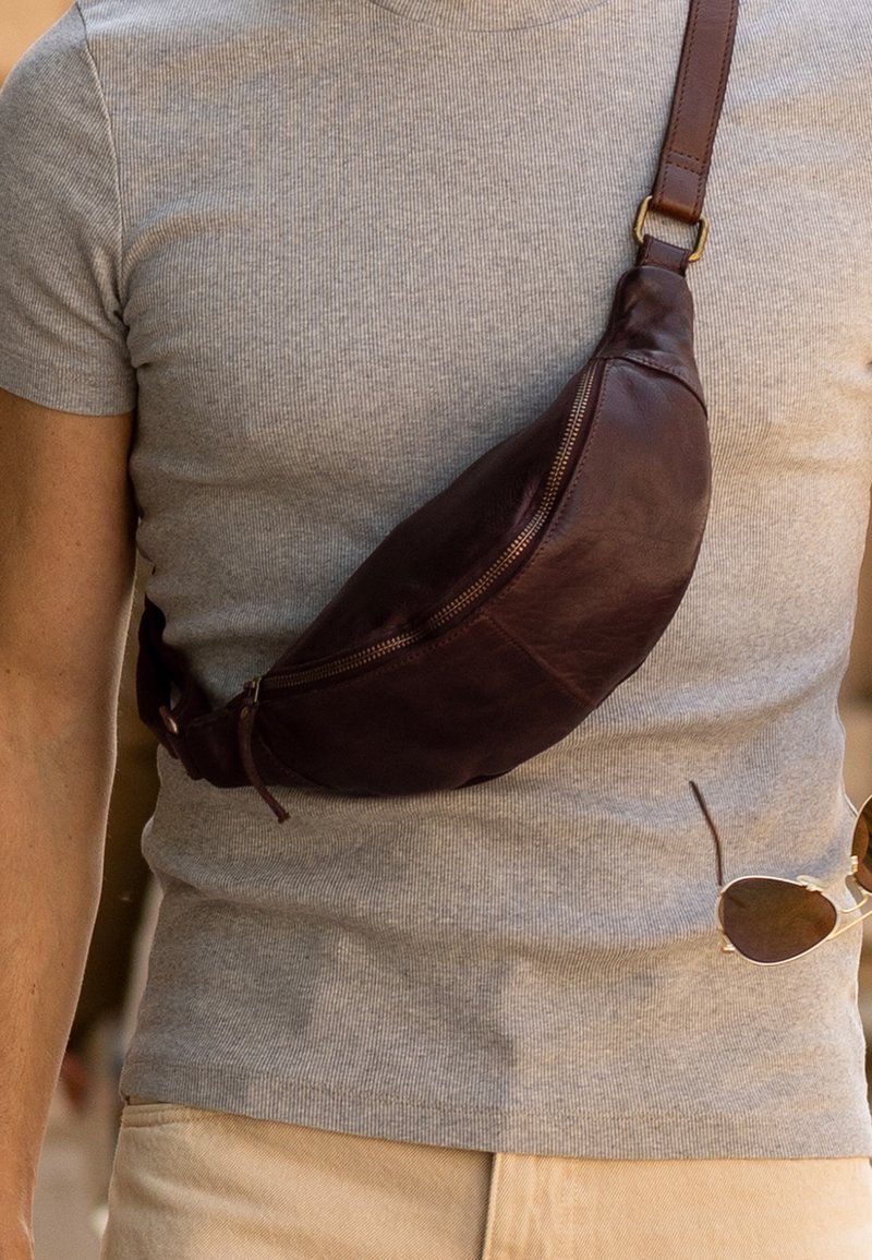 Mand iført grå ribbet skjorte med brun læder crossbody taske og holder runde solbriller i hånden.