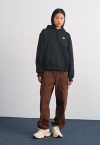 Sweat-shirt à capuche noire avec une poche avant, associé à un pantalon cargo marron et des baskets crème. Tissu lisse, coupe décontractée, logo visible.