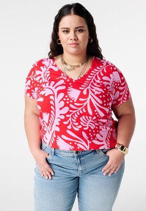 Vrouw met lang donker haar draagt een roodroze bloemetjespatroon blouse, blauwe spijkerbroek, gouden oorringen, gelaagde kettingen en een gouden polshorloge.