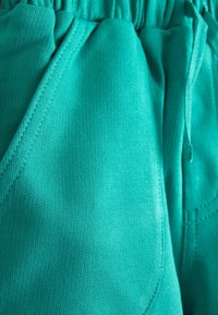 Prim-plan cu pantaloni din material turcoaz, cu cusături vizibile ale buzunarelor, talie elastică și șnur reglabil în partea dreaptă.