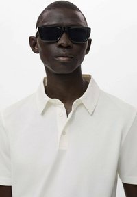 White polo shirt with a collar, featuring a button placket. Worn with black sunglasses. Smooth texture and tailored fit.