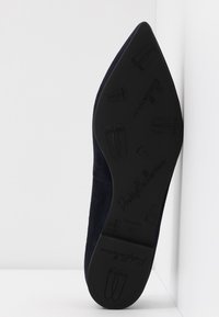Shoe flat em camurça azul-marinho com bico pontudo e sola de borracha texturizada. Possui um logotipo da marca em relevo e silhuetas de sapatilhas na parte inferior.